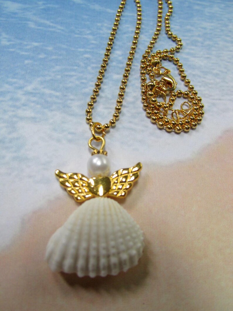 May include: A gold necklace with a seashell angel pendant. The pendant features a white seashell body, gold wings, and a pearl head. The necklace chain is made of small gold beads, and the clasp is visible.