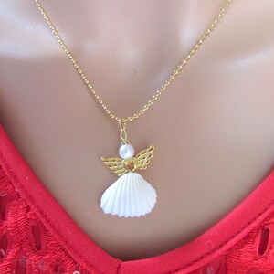 May include: A gold-coloured necklace with an angel pendant. The pendant has a white seashell body, gold-coloured wings, and a pearl head. The necklace is shown against a red lace top.