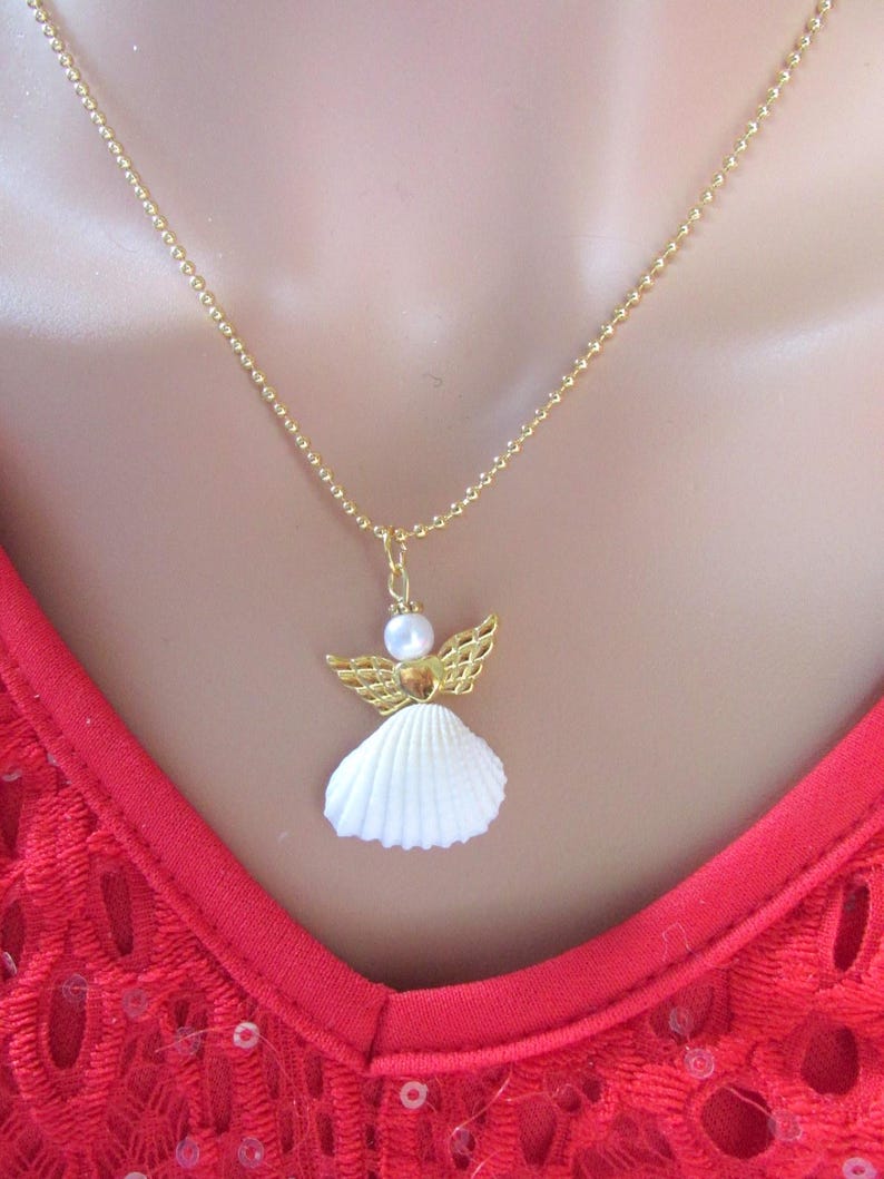 May include: A gold-coloured necklace with an angel pendant. The pendant has a white seashell body, gold-coloured wings, and a pearl head. The necklace is shown against a red lace top.