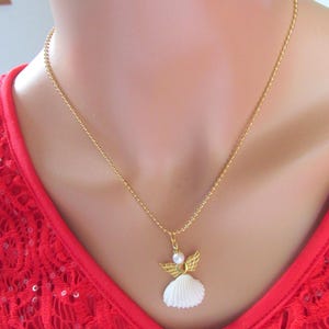 May include: A gold necklace with an angel pendant. The pendant has a white seashell body, gold wings, and a pearl head. The necklace is set against a red background. The chain is a delicate, beaded style.