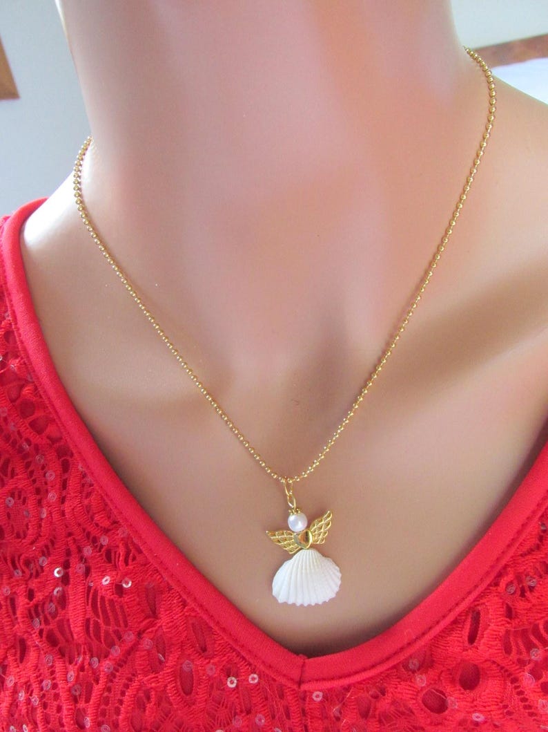 May include: A gold necklace with an angel pendant. The pendant has a white seashell body, gold wings, and a pearl head. The necklace is set against a red background. The chain is a delicate, beaded style.
