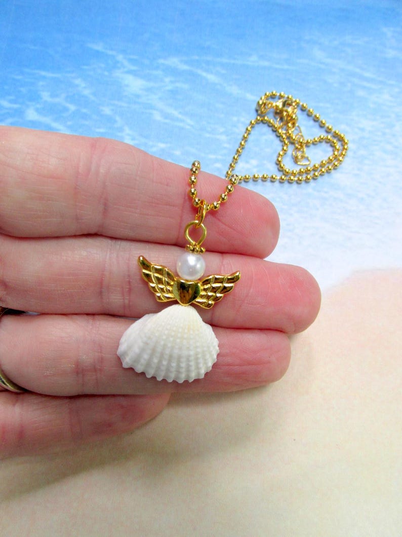 May include: A gold necklace with a seashell angel pendant. The pendant features a white seashell body, gold wings, and a pearl head. The necklace chain is gold.