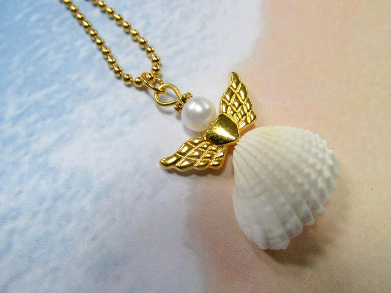 May include: A gold-coloured necklace with an angel pendant. The pendant has gold wings, a pearl, and a white seashell body. The necklace chain is made of small, gold-coloured beads.