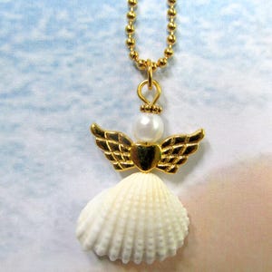 May include: A gold angel pendant necklace. The pendant has a white seashell skirt, gold wings, a pearl head, and a gold heart. The necklace chain is a delicate gold ball chain.