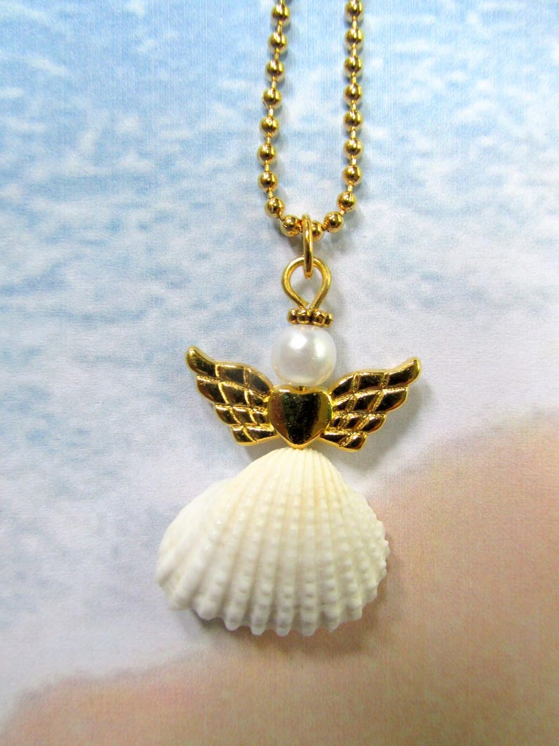 May include: A gold angel pendant necklace. The pendant has a white seashell skirt, gold wings, a pearl head, and a gold heart. The necklace chain is a delicate gold ball chain.