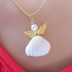 May include: A gold necklace with an angel pendant. The angel pendant has a white seashell skirt, gold filigree wings, a pearl head, and a gold heart-shaped body. The necklace chain is gold.