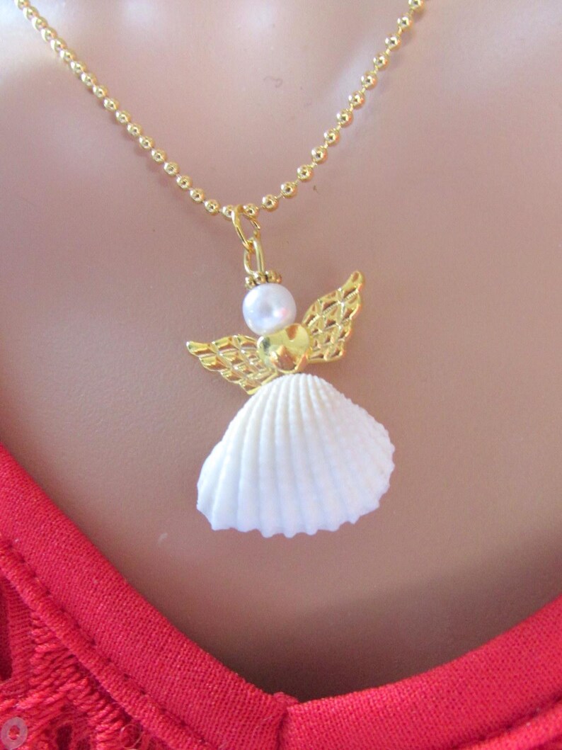 May include: A gold necklace with an angel pendant. The angel pendant has a white seashell skirt, gold filigree wings, a pearl head, and a gold heart-shaped body. The necklace chain is gold.