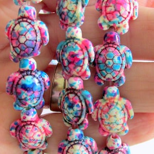 May include: Three strands of turtle-shaped beads featuring a speckled pattern in vibrant shades of pink, blue, yellow, and white. Each bead is carved with a detailed shell design and strung on a clear cord, creating a colorful and textured appearance.