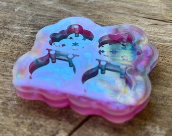 Könnte beinhalten: Eine wolkenförmige Silikonform mit zwei Hohlräumen. Die Form hat einen Farbverlauf aus Rosa und schillernden Farben mit einem transparenten Innenraum. Jeder Hohlraum hat ein Wolken- und Sterndesign mit kleinen Stiften zum Aufhängen.