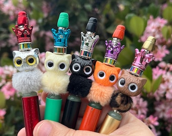 May include: Six colorful pens with cat-shaped tops and crowns. Each pen has a different colored crown and a different colored fur pom-pom.