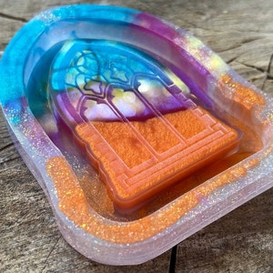May include: A decorative resin tray with a stained glass window design. The tray features a vibrant mix of colors, including blue, purple, and orange, with glitter accents. The window design is orange and has a detailed outline.
