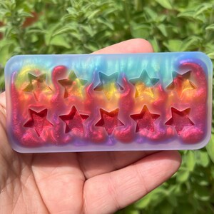 May include: A rectangular resin mould with five star-shaped cutouts. The resin is multi-coloured, transitioning from blue to yellow to red, with an iridescent shimmer. The mould is held in a hand against a backdrop of green foliage.