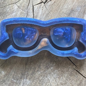 以下が含まれることがあります： サングラスの形をした青いグリッターレジンモールド。このモールドは、透明なレジン製のサングラスを作るように設計されています。