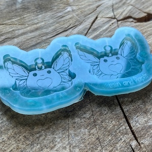 May include: Clear silicone mold with two cavities shaped like a fairy with wings and a heart. The mold is for making resin crafts and has the text "Resin and Paper" on the bottom.