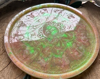 May include: A round, clear resin tray with a gold glitter rim. The tray features a white design of the zodiac signs and a crescent moon. The background is a light pink with green glitter.