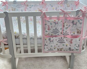 Op de afbeelding: Witte wieg met een roze-witte geprinte wiegbumper en een roze-witte geprinte hangende organizer met meerdere vakken. De organizer heeft een hert- en bloemenprint.