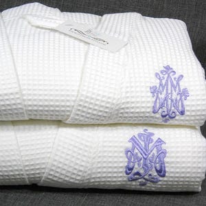May include: Two white waffle weave bathrobes with purple embroidered monograms. The top robe has the monogram "MMW" and the bottom robe has the monogram "NYW".