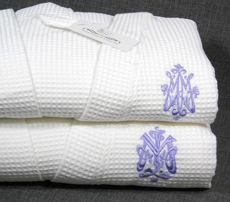 May include: Two white waffle weave bathrobes with purple embroidered monograms. The top robe has the monogram "MMW" and the bottom robe has the monogram "NYW".