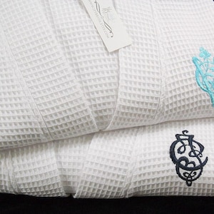 May include: Two white waffle weave bathrobes with embroidered initials. The top robe has a turquoise blue monogram and the bottom robe has a black monogram.