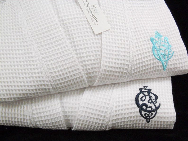 May include: Two white waffle weave bathrobes with embroidered initials. The top robe has a turquoise blue monogram and the bottom robe has a black monogram.