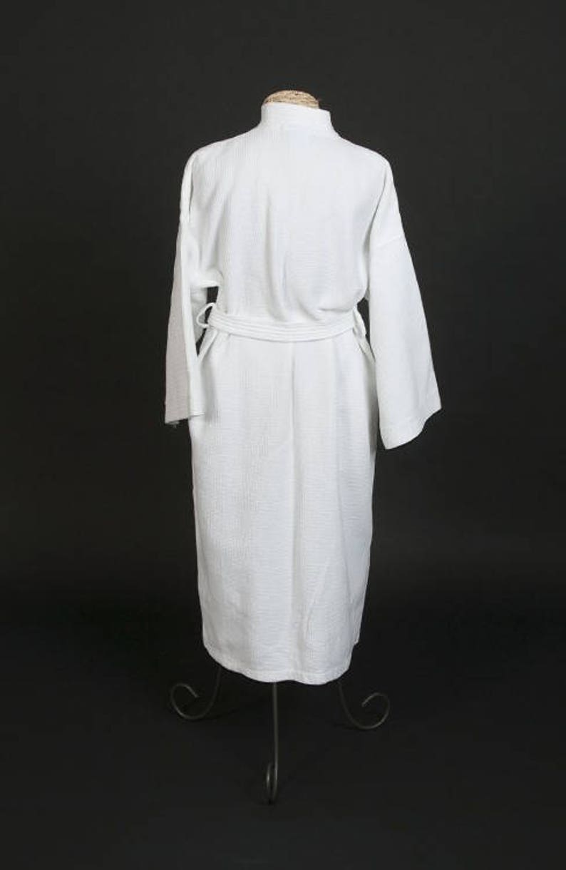 May include: A white waffle weave bathrobe with a self-tie belt. The robe is hanging on a stand against a black background.