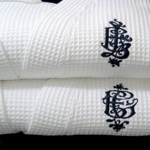 May include: Two white waffle weave bathrobes with a black embroidered monogram of the letters "E.B." on each robe.