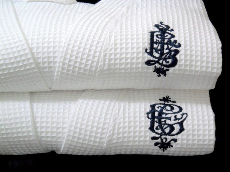 May include: Two white waffle weave bathrobes with a black embroidered monogram of the letters "E.B." on each robe.
