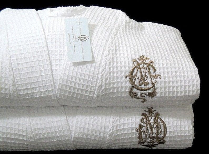 May include: White waffle weave bathrobe with a gold embroidered monogram. The robe is folded in half and the monogram is visible on both sides.