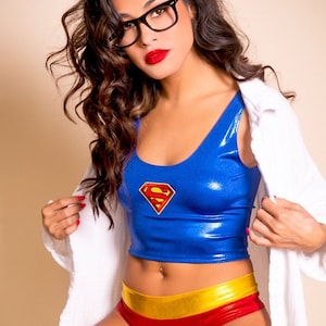 May include: A woman wearing a blue and red superhero costume with a gold belt and a Superman logo on the chest. She is wearing black framed glasses and has long brown hair. She is holding a white shirt in her hands.