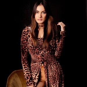 Op de afbeelding: Een vrouw draagt een outfit met luipaardprint, bestaande uit een jurk met lange mouwen en een korte broek. De jurk heeft een split aan de voorkant, waardoor de korte broek zichtbaar is. Ze draagt een zwarte choker. De achtergrond is donker.