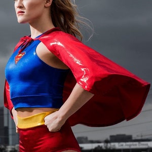 K&ouml;nnte beinhalten: Eine Person in einem Superhelden-Kost&uuml;m steht vor einem bew&ouml;lkten Himmel mit einer Skyline im Hintergrund. Das Kost&uuml;m umfasst ein blaues Oberteil, rote Shorts, einen gelben G&uuml;rtel und einen roten Umhang.