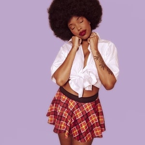 K&ouml;nnte beinhalten: Eine Frau mit Afrofrisur tr&auml;gt ein wei&szlig;es Hemd mit Kn&ouml;pfen, das in der Taille gebunden ist, und einen rot-schwarzen karierten Rock.