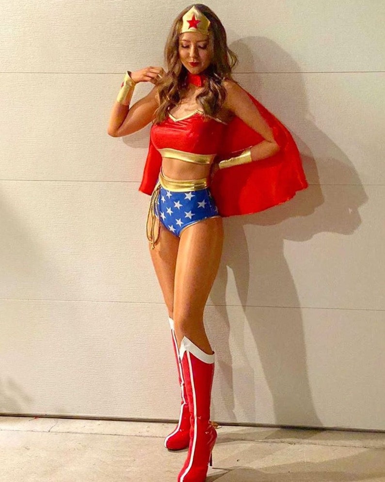 May include: A woman dressed as Wonder Woman for Halloween. She is wearing a red and gold bustier, blue and gold shorts with white stars, red boots with white accents, and a red cape. She is also wearing a gold headband with a red star.