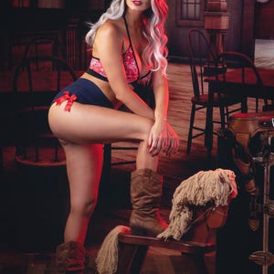 May include: A woman wearing a red and white patterned bikini top and blue denim shorts with a red bow. She is wearing a brown cowboy hat and brown cowboy boots. She is sitting on a wooden rocking horse with a white mane.