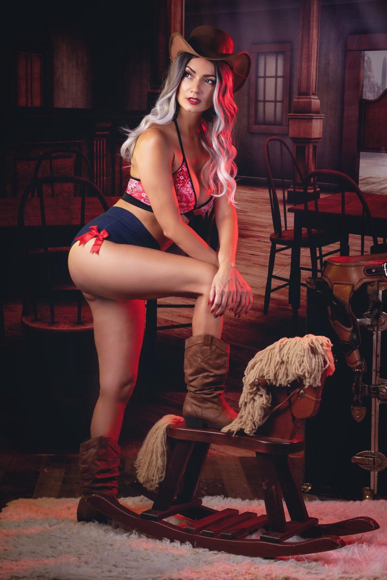 May include: A woman wearing a red and white patterned bikini top and blue denim shorts with a red bow. She is wearing a brown cowboy hat and brown cowboy boots. She is sitting on a wooden rocking horse with a white mane.