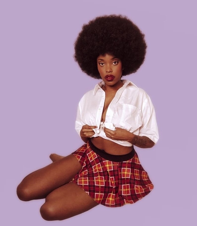 K&ouml;nnte beinhalten: Eine Frau mit Afro-Frisur tr&auml;gt ein wei&szlig;es Hemd mit Kn&ouml;pfen und einen rot-schwarzen karierten Rock. Sie sitzt auf einem lila Hintergrund.