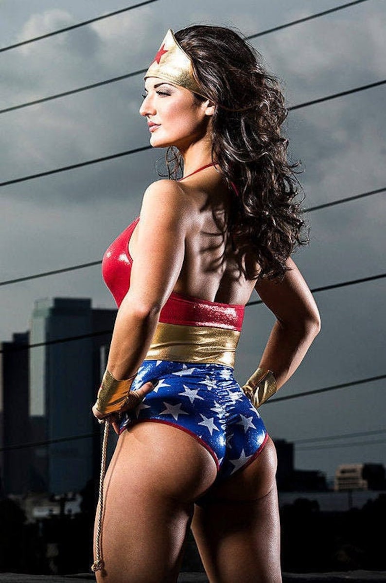 May include: A woman dressed as Wonder Woman, wearing a red, gold and blue costume with a star-spangled design. She is standing with her back to the camera, looking over her shoulder.