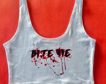 May include: White crop top with the text "BITE ME" in black and red, with red splatter details. The top has a scoop neck and tank top straps. The background is a solid red.