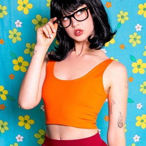 Puede incluir: Un top corto naranja y una falda burdeos se usan con una peluca negra y gafas. El fondo es un estampado floral azul. El atuendo recuerda a un personaje de dibujos animados.