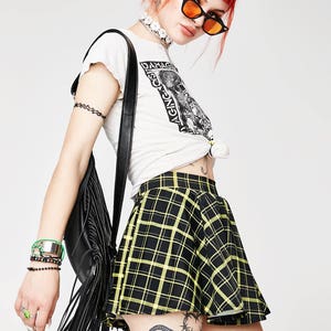 May include: A black and yellow plaid mini skirt with a high waist and a flared silhouette. The skirt is worn by a person with red hair and a white t-shirt with the text "Damaged Goods Aging Against" printed on it.