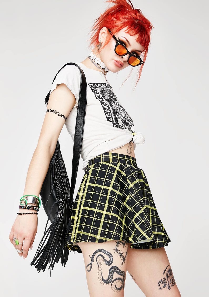 May include: A black and yellow plaid mini skirt with a high waist and a flared silhouette. The skirt is worn by a person with red hair and a white t-shirt with the text "Damaged Goods Aging Against" printed on it.