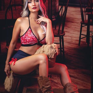 May include: A woman wearing a red and white bandana print bra top and denim shorts sits on a rocking horse. She is wearing a brown cowboy hat and brown cowboy boots. The background is a rustic saloon setting.