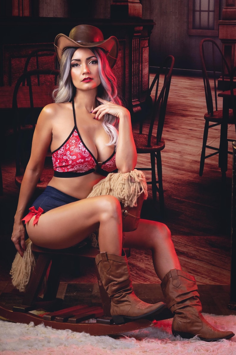 May include: A woman wearing a red and white bandana print bra top and denim shorts sits on a rocking horse. She is wearing a brown cowboy hat and brown cowboy boots. The background is a rustic saloon setting.