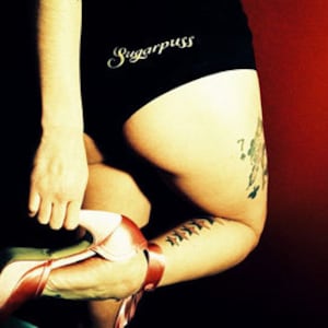 May include: A woman wearing black underwear with the text "Sugarpuss" on the waistband. She has a tattoo on her leg with playing cards and green leaves. She is wearing a pink high heel shoe.