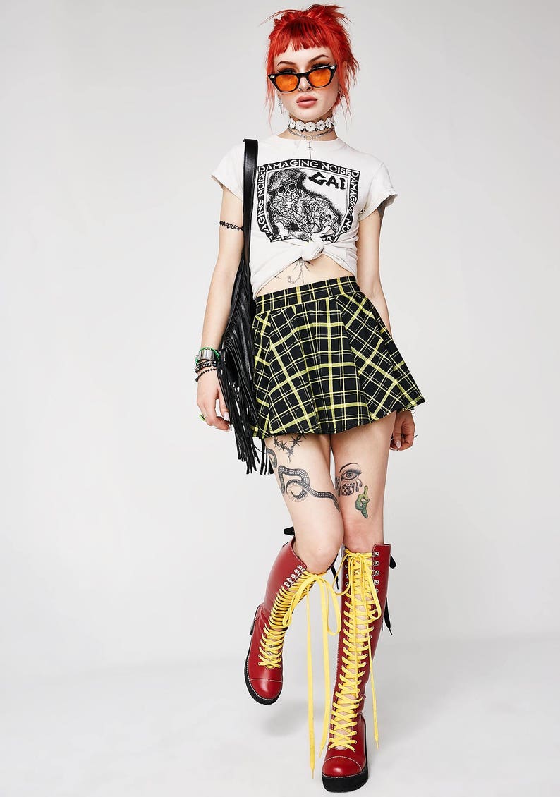 May include: A woman wearing a white t-shirt with a black and white graphic print that says "Damaging Noise", a black and yellow plaid skirt, red boots with yellow laces, and orange sunglasses.