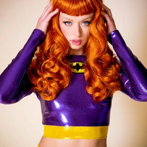 May include: A purple and yellow superhero costume with a long-sleeved top and a skirt. The top features a yellow bat symbol on the chest. The model has bright red hair and is wearing the costume.