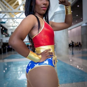 May include: A woman dressed as Wonder Woman, wearing a red, gold and blue costume with a star-spangled design. She is posing with her arm raised, showing off her bicep.