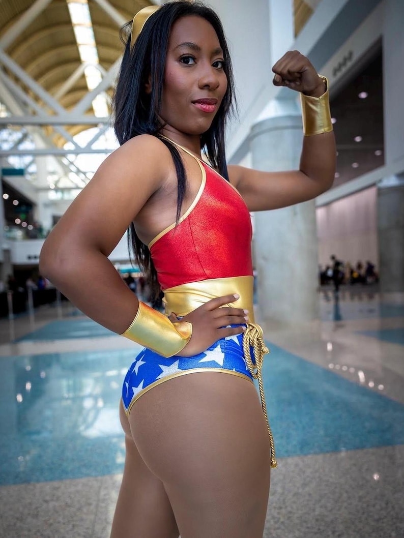 May include: A woman dressed as Wonder Woman, wearing a red, gold and blue costume with a star-spangled design. She is posing with her arm raised, showing off her bicep.