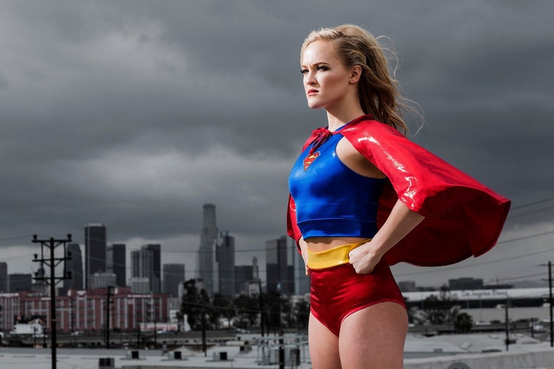 K&ouml;nnte beinhalten: Eine Person in einem Superhelden-Kost&uuml;m steht vor einem bew&ouml;lkten Himmel mit einer Skyline im Hintergrund. Das Kost&uuml;m umfasst ein blaues Oberteil, rote Shorts, einen gelben G&uuml;rtel und einen roten Umhang.