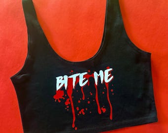 May include: Black crop top with the text "BITE ME" in white, with red dripping blood effect. The top has a scoop neck and wide straps. The background is a solid red.
